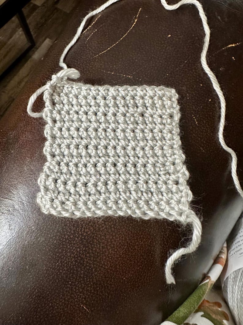 May include: A small, square swatch of light gray crochet fabric. The stitches are tightly woven, creating a textured surface. White yarn extends from the top and right side of the swatch, indicating the beginning and end of the work. The swatch is on a brown surface.