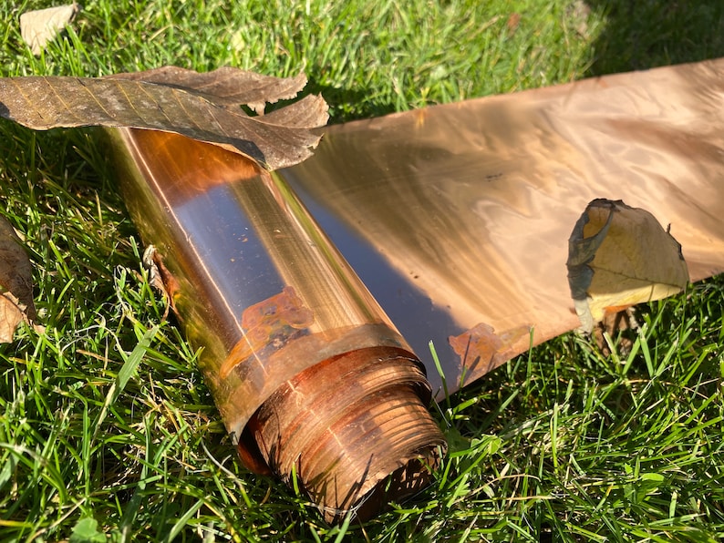 May include: A rolled sheet of copper foil with a shiny, metallic finish. The foil is lying on a bed of green grass with some dried leaves.