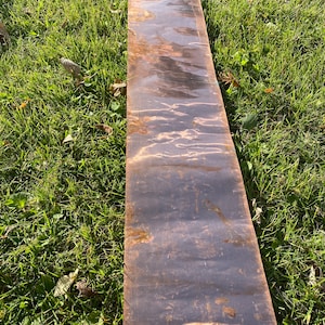 May include: A long, narrow sheet of copper with a dark brown patina. The copper sheet has a slightly textured surface and is lying on a bed of green grass.