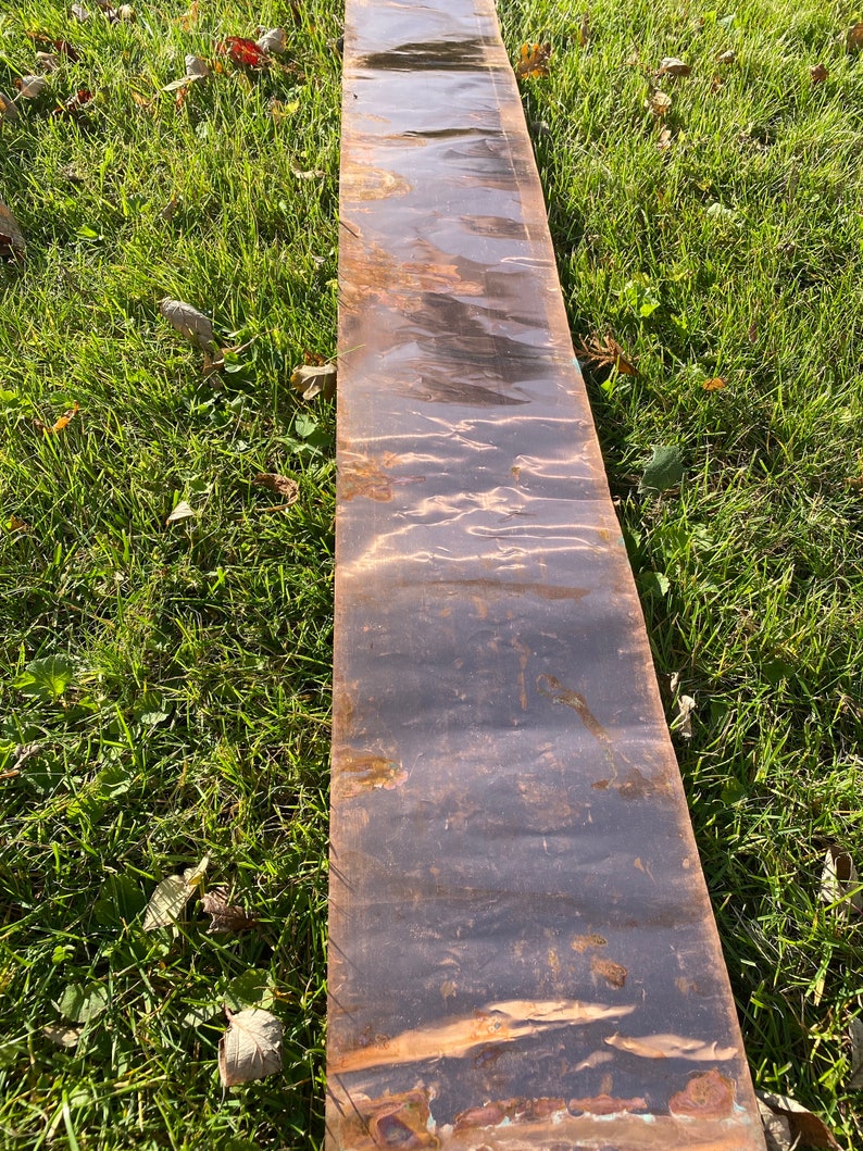 May include: A long, narrow sheet of copper with a dark brown patina. The copper sheet has a slightly textured surface and is lying on a bed of green grass.