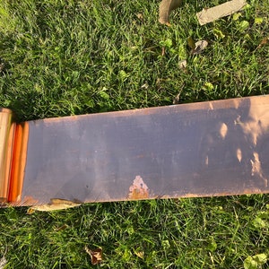May include: A rolled up sheet of copper flashing, showing the shiny copper surface and a slight amount of rust. The flashing is lying on a bed of green grass.