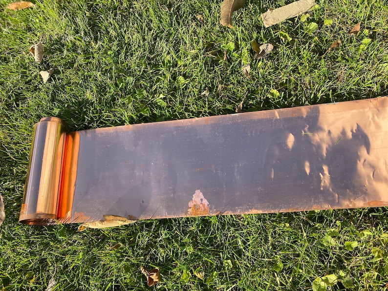 May include: A rolled up sheet of copper flashing, showing the shiny copper surface and a slight amount of rust. The flashing is lying on a bed of green grass.