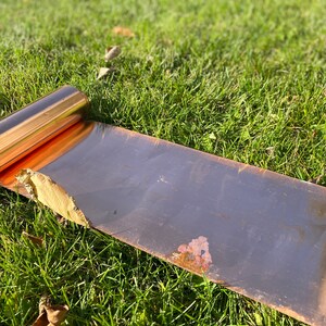 May include: A roll of copper sheet metal lying on green grass. The sheet metal is partially unrolled, revealing the shiny copper surface.