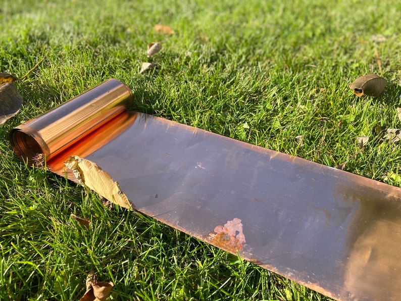 May include: A roll of copper sheet metal lying on green grass. The sheet metal is partially unrolled, revealing the shiny copper surface.