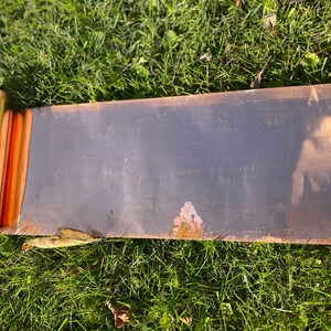 May include: A rolled copper sheet metal panel, with a slight bend in the middle, lying on a bed of green grass.