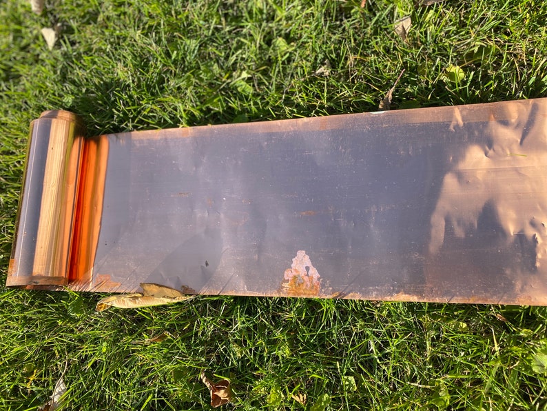 May include: A rolled copper sheet metal panel, with a slight bend in the middle, lying on a bed of green grass.