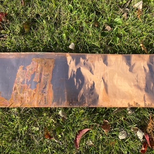 May include: A long, thin metal sheet with a dark brown patina on one side and a lighter brown patina on the other side. The metal sheet has a textured surface.