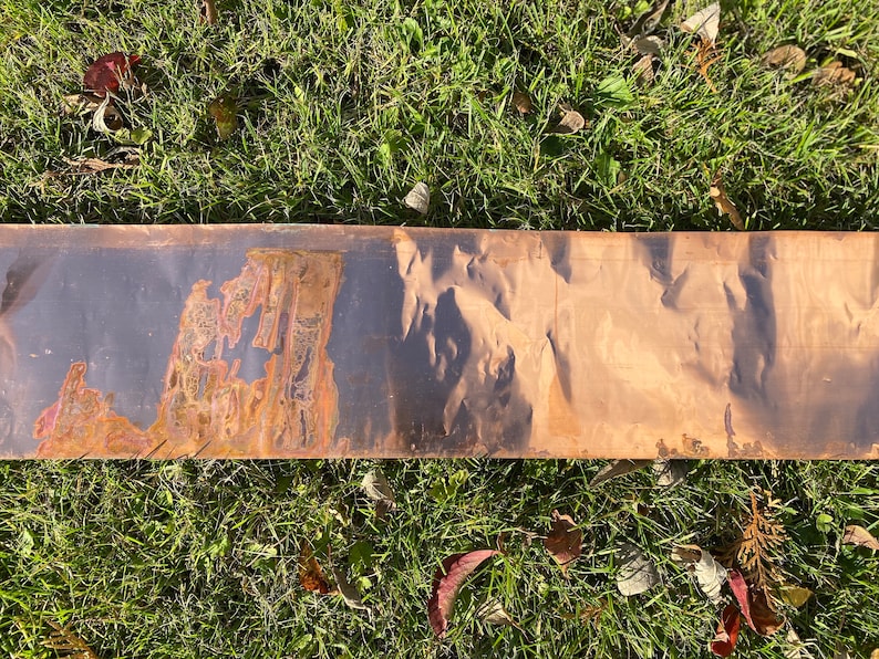 May include: A long, thin metal sheet with a dark brown patina on one side and a lighter brown patina on the other side. The metal sheet has a textured surface.