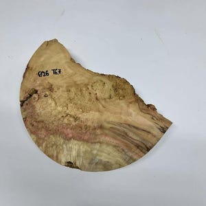 Puede incluir: Una losa de madera natural de forma irregular con borde sin tratar. La madera presenta una mezcla de tonos marrón claro, rosa y marrón oscuro, con vetas y nudos visibles. El texto "026 76.7" está escrito en la superficie.