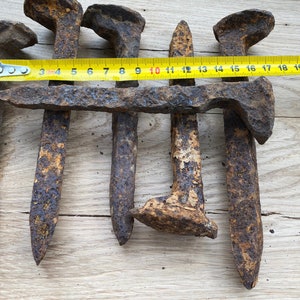May include: Close-up of six rusty railway spikes, some with square heads and some with pointed heads. The spikes are arranged in a row and are measured with a yellow tape measure that reads "500 34 5 6 7 8 9 10 11 12 13 14 15 16 17 18 19 20 21 22".