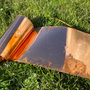 May include: A roll of copper sheet metal with a patina finish. The metal is rolled up and partially unrolled, showing the shiny copper underneath. The metal is lying on a bed of green grass.