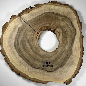 May include: A large, round wooden slice with a hole in the centre. The wood has a natural brown and light brown colour with a unique grain pattern. The slice is labelled "N 253 35x36x2.8".