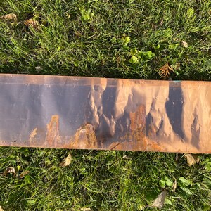 May include: A long, thin, rectangular piece of metal with a brown, rust-like patina. The metal is slightly bent and has a textured surface.