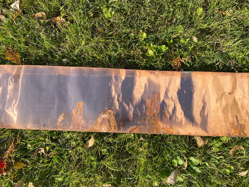 May include: A long, thin, rectangular piece of metal with a brown, rust-like patina. The metal is slightly bent and has a textured surface.