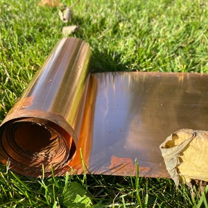 May include: A roll of copper sheet metal, partially unrolled, lying on green grass. The copper has a shiny, metallic finish and shows signs of oxidation.