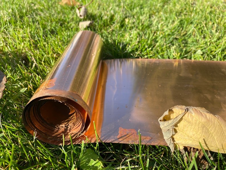 May include: A roll of copper sheet metal, partially unrolled, lying on green grass. The copper has a shiny, metallic finish and shows signs of oxidation.