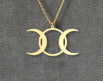May include: A gold-colored necklace featuring a triple moon pendant. The pendant consists of three connected crescent moons, symbolizing the phases of the moon. The necklace hangs on a delicate gold chain, set against a gray background.