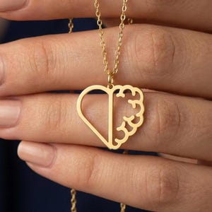 May include: A gold-toned necklace featuring a pendant shaped like a heart. The left side of the heart is a simple smooth outline, while the right side features a detailed, textured design resembling the intricate folds of a human brain.