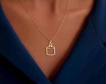 May include: A gold-colored necklace featuring a pendant shaped like a candle. The candle pendant has a rectangular body with a flame at the top. The necklace is worn against a dark blue garment.