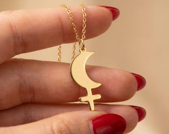 May include: A gold-colored necklace featuring a pendant with a crescent moon and a cross symbol. The pendant hangs from a delicate chain. The necklace is held against a neutral background.