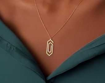 May include: A gold-colored pendant necklace featuring a geometric paperclip design. The pendant hangs from a delicate chain. The necklace is displayed against a teal-colored top.