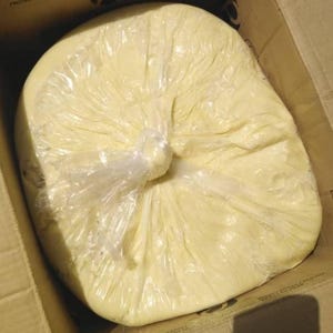 May include: A large, pale yellow block, possibly food, wrapped in clear plastic. The wrapped item is inside a cardboard box. The plastic is tied at the top, and the item fills most of the box.