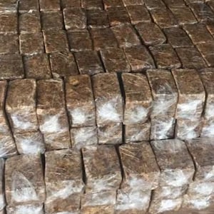 以下が含まれることがあります： 透明なプラスチックで包まれた茶色の長方形のブロックの山。ブロックはきれいに並べて積み重ねられています。