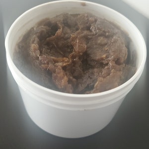 Puede incluir: Un recipiente de pl&aacute;stico blanco lleno de una sustancia espesa y texturizada de color marr&oacute;n oscuro. La sustancia parece ser un exfoliante corporal o jab&oacute;n hecho en casa.
