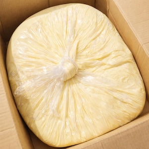May include: A large, round block of pale yellow, creamy substance encased in a clear, thin plastic film. The plastic is gathered and twisted into a knot at the centre of the top surface, creating radiating folds across the smooth, solid mass.