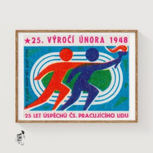 May include: A framed vintage matchbox with a design of two running figures, one red and one blue, carrying a torch. The text reads "25. VÝROČÍ ÚNORA 1948" and "25 LET ÚSPĚCHŮ ČS. PRACUJÍCÍHO LIDU" on a blue background. The matchbox is in a wooden frame.