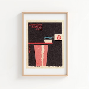 Pode incluir: Uma impressão vintage emoldurada com uma escova de dentes, pasta de dentes e um copo. A obra de arte usa vermelho, azul e branco sobre um fundo preto. O texto "ZABRAŇUJE ZUBNÍMU KAZU" é exibido na parte superior.