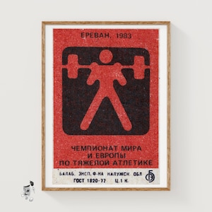 May include: Framed vintage poster with a red background and black graphic of a weightlifter. The poster includes text in Russian, with the year 1983 visible at the top. The poster is in a wooden frame.