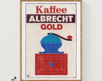 May include: A framed vintage-style print featuring the text Kaffee ALBRECHT GOLD in red and dark brown. Below the text is a graphic illustration of a blue coffee grinder with a red base and a dark brown handle, all set on a textured off-white background.
