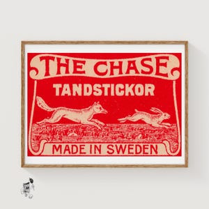 May include: A framed vintage print with a red background and the words "THE CHASE" and "TANDSTICKOR" in cream-colored text. The print features a fox chasing a rabbit. The words "MADE IN SWEDEN" are at the bottom.