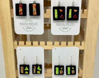 May include: A display of eight pairs of handmade glass earrings. The earrings are square shaped and feature geometric designs in various colors, including red, green, blue, yellow, and black. The earrings are displayed on a wooden shelf with the text "PEACEFUL PURSUITS Studio" on each card.