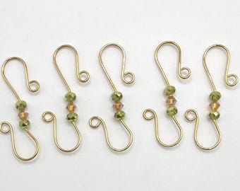 May include: Five gold-toned metal S-hooks, each featuring a central decorative segment composed of two small olive-green faceted beads flanking a single amber-colored faceted bead, arranged in a row against a plain white background.