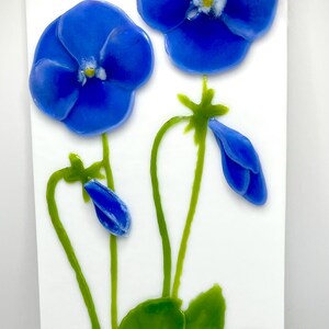 May include: A vibrant artwork featuring two large, blue flower blossoms with yellow centers, alongside green stems, leaves, and a bud. The flowers are set against a white background, creating a striking contrast. The artwork evokes a sense of nature and botanical beauty.