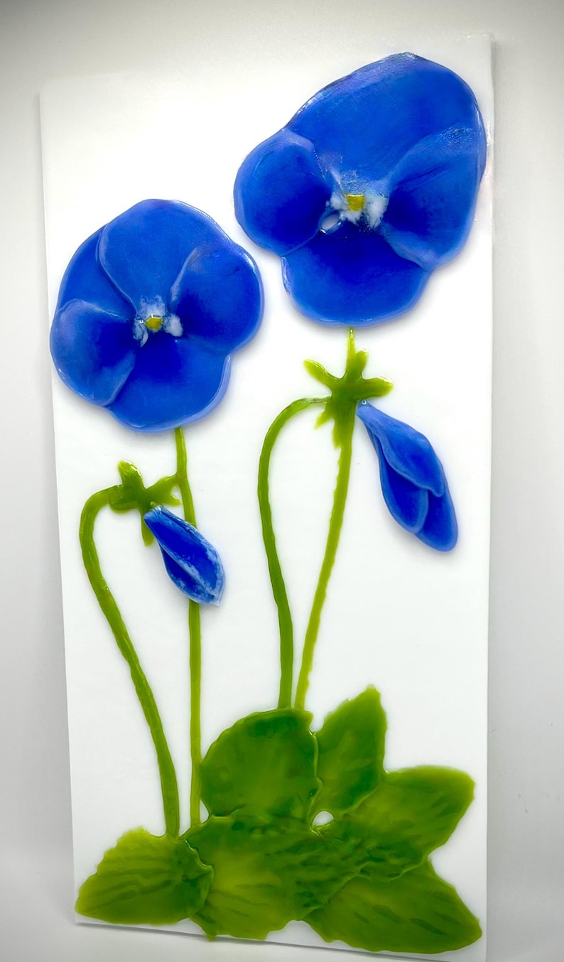 May include: A vibrant artwork featuring two large, blue flower blossoms with yellow centers, alongside green stems, leaves, and a bud. The flowers are set against a white background, creating a striking contrast. The artwork evokes a sense of nature and botanical beauty.