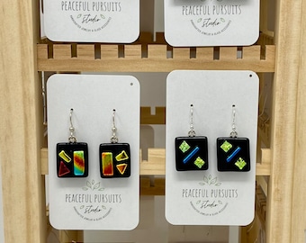 May include: Six pairs of black square earrings with colorful geometric designs. The earrings are displayed on a wooden shelf with white cards that read "PEACEFUL PURSUITS Studio".