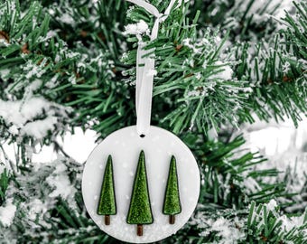 May include: A white, round Christmas ornament with three green glittery Christmas trees. The ornament hangs from a white ribbon against a backdrop of a snowy green Christmas tree. The trees have brown trunks.