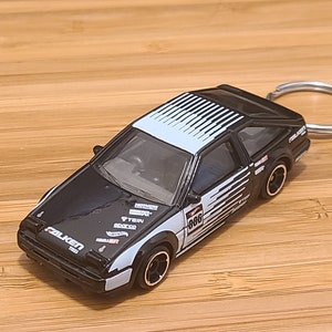 May include: Black and white toy car keychain with a white and black racing stripe design. The car has the number "886" on the side and the word "Falken" on the front.