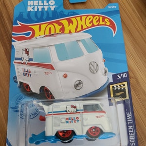 May include: White Hot Wheels toy car with a Hello Kitty design. The car is a Volkswagen van with a blue and red stripe and a Hello Kitty logo on the side. The car is on a blue background with the words "Hot Wheels" and "Hello Kitty" printed on it.