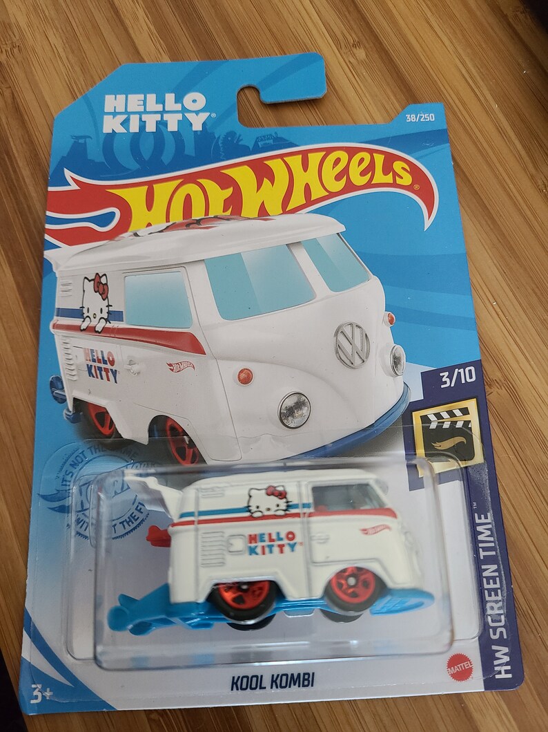 May include: White Hot Wheels toy car with a Hello Kitty design. The car is a Volkswagen van with a blue and red stripe and a Hello Kitty logo on the side. The car is on a blue background with the words "Hot Wheels" and "Hello Kitty" printed on it.