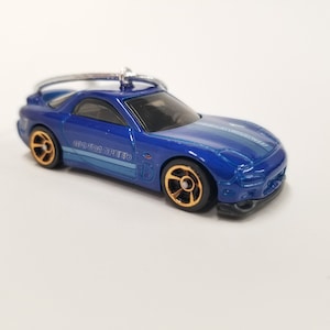 May include: A blue Mazda RX-7 toy car with gold wheels and "Mazda Speed" written on the side. The car is a die-cast model and is a popular collectible.