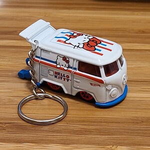 May include: White Hello Kitty van keychain with red and blue stripes, a Hello Kitty face on the roof, and a Hello Kitty logo on the side. The keychain has a silver metal ring.