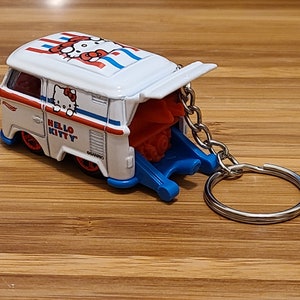 May include: White Hello Kitty van keychain with a red and blue design. The van has a pop-up back and a silver chain.