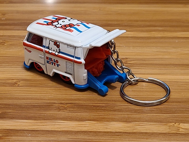 May include: White Hello Kitty van keychain with a red and blue design. The van has a pop-up back and a silver chain.