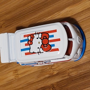 May include: A white toy van with a red, white, and blue striped roof. The roof features a Hello Kitty graphic with a red bow.