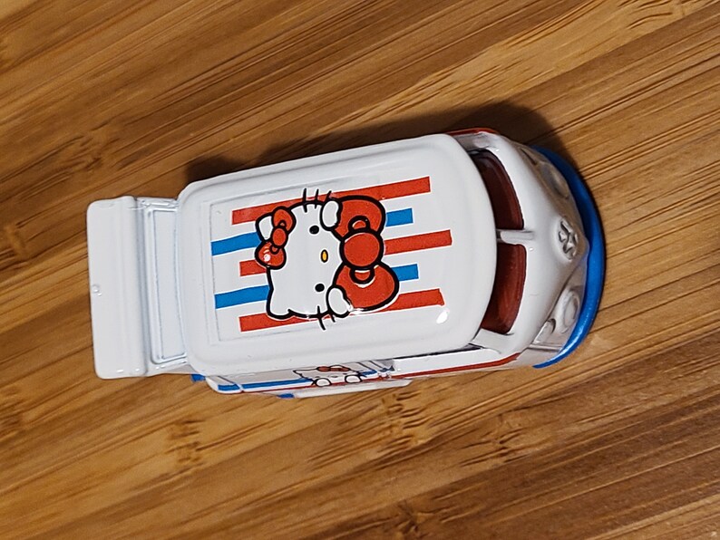 May include: A white toy van with a red, white, and blue striped roof. The roof features a Hello Kitty graphic with a red bow.