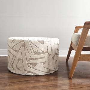 Puede incluir: Un otom&aacute;n cil&iacute;ndrico con tela color crema con un patr&oacute;n abstracto repetido en un tono marr&oacute;n claro. El otom&aacute;n est&aacute; sobre un piso de madera, con una silla de madera visible en el fondo.